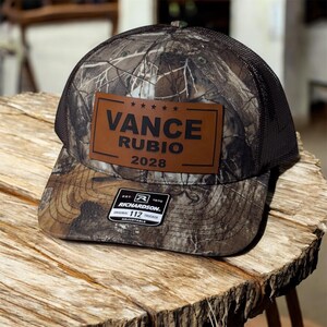 May include: A camouflage trucker hat with a brown leather patch displaying the text "VANCE RUBIO 2028". The hat has a black mesh back and a curved brim. The Richardson 112 Trucker hat is adjustable.