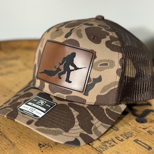 May include: A brown and tan camouflage trucker hat with a brown leather patch featuring a silhouette of a Bigfoot holding a rifle and a deer. The hat has a brown mesh back and a black adjustable strap with a white label that reads "Richardson 112 Trucker Adjustable".