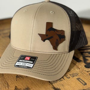 May include: A tan and black trucker hat with a brown leather Texas state outline patch. The patch features a star, a longhorn steer, and a pistol. The hat has a black mesh back and a brown adjustable strap with the text "Richardson 112 Original Trucker Adjustable" on a black label.