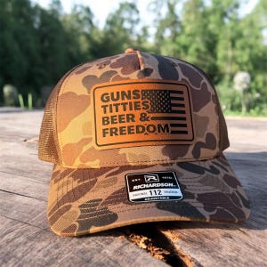 May include: Camouflage trucker hat with a brown mesh back and a brown patch featuring the text "GUNS, TITTIES, BEER & FREEDOM" and an American flag design. The hat is adjustable and has a Richardson 112 Trucker label.