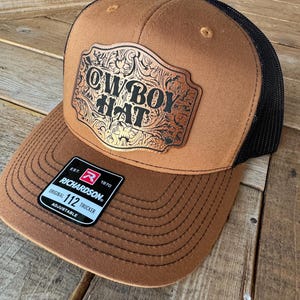 May include: A brown and black trucker hat with the words "COWBOY HAT" in a decorative gold-colored emblem. The hat has a mesh back and a curved brim. The Richardson brand tag is visible.