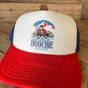 May include: A red, white, and blue trucker hat with a graphic of a man sitting in water under an American flag design. The text on the hat reads "Hotter than a Hoochie Coochie."