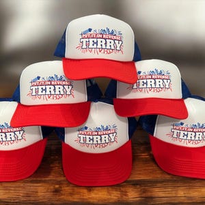 May include: A stack of six trucker hats with white fronts, red brims, and blue mesh backs. The hats feature the text "PUT IT IN REVERSE TERRY" in a red and blue design.