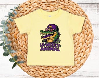 May include: A light yellow t-shirt featuring a cartoon alligator wearing a purple hat and sunglasses. The alligator is green and yellow with a purple mouth. The text "LA-WEEZY ANIMAL" is printed below the alligator in purple and white.