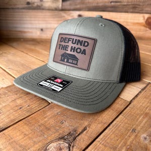 May include: A green and black trucker hat with a brown leather patch that says "Defund the HOA".