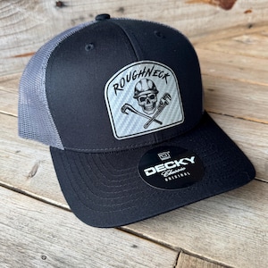 May include: Black and gray trucker hat with a curved brim. The hat features a patch with the word "ROUGHNECK" above a skull wearing a hard hat and crossed wrenches. The hat is made by Decky.