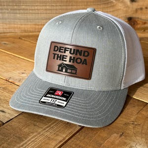 May include: Gray and white trucker hat with a brown leather patch that says "Defund the HOA" with a house icon. The hat is a Richardson 112 Original Trucker.