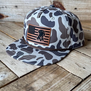 May include: A camouflage-patterned baseball cap with a brown leather patch featuring an American flag design and a dog silhouette. The cap has a brown rope detail along the brim and a button on top.