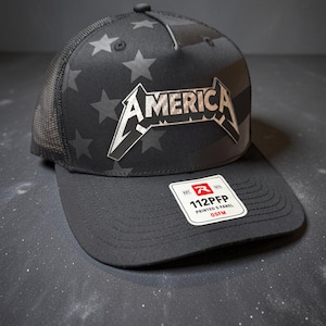 May include: A black five-panel trucker hat featuring a tonal star and stripe pattern on the front panels. The word AMERICA is printed in a metallic silver font across the front. A white sticker on the brim reads R EST 1970 112PFP PRINTED 5 PANEL OSFM.