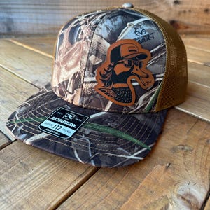 May include: A brown and tan camouflage trucker hat with a brown leather patch featuring a cartoon duck wearing sunglasses. The hat has a mesh back and the text "REALTREE MAX-1" is visible on the front. The hat is made by Richardson 112.