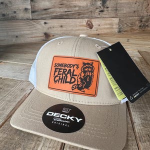 May include: Tan and white trucker hat with a patch that reads "SOMEBODY'S FERAL CHILD" and features a raccoon illustration. The hat has a curved brim and a mesh back. A black tag hangs from the hat.