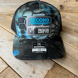May include: A black and blue camouflage trucker hat with a patch that says "Second Amendment Protecting Freedom Since 1791". The patch features a black and white illustration of a rifle. The hat is adjustable and has a Richardson 112 label.
