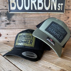 May include: Two baseball caps with the text "OPERATION MIDNIGHT HAMMER" on a patch. One cap is black with a yellow brim, the other is olive green. A sign that reads "BOURBON ST" is in the background.