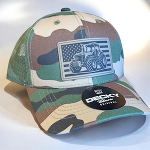 May include: Camouflage baseball cap with a tractor and American flag patch. The cap features a curved brim, a mesh back, and a button on top. The patch has a detailed tractor design. The hat is a Decky Classic Original.