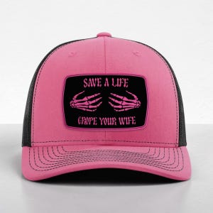 May include: A bright pink trucker hat with a black mesh back and a rectangular patch on the front. The patch is black with a pink border and features two pink skeleton hands and the text "SAVE A LIFE GROPE YOUR WIFE" in pink lettering.