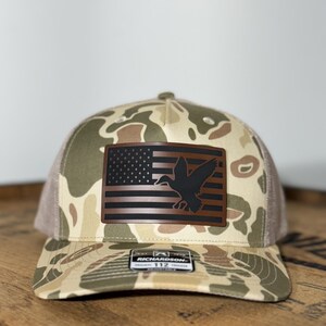 May include: A camouflage trucker hat with a brown leather patch featuring a black silhouette of a duck flying over an American flag. The hat is made by Richardson 112.