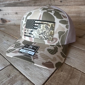 May include: Camouflage pattern trucker hat with a patch featuring a distressed American flag and a rattlesnake. The hat has a curved brim, a mesh back, and the text "DON'T TREAD ON ME". The Richardson 112 Trucker hat is adjustable.