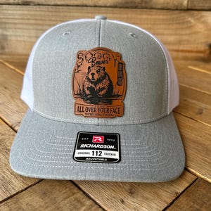 May include: Gray and white trucker hat with a brown leather patch featuring a beaver illustration and the text "Beaver BBQ". The hat also has the text "ALL OVER YOUR FACE YOU'RE EATING IT WRONG". Adjustable fit.