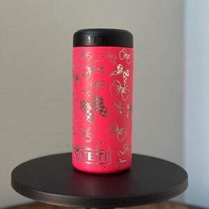 以下が含まれることがあります： 銀色のボウのパターンが施されたピンクのYETIランブラー。 YETIのロゴがタンブラーの側面に見えます。