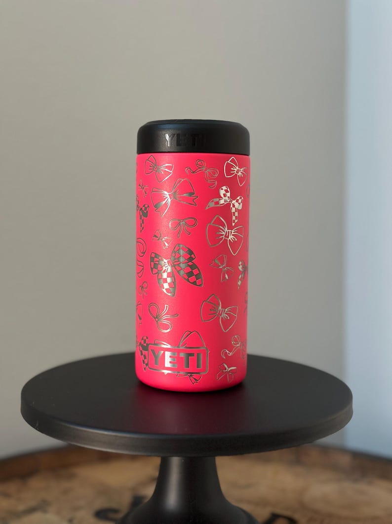 以下が含まれることがあります： 銀色のボウのパターンが施されたピンクのYETIランブラー。 YETIのロゴがタンブラーの側面に見えます。