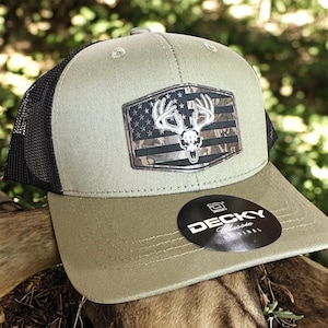 May include: A green and black trucker hat with a patch featuring a deer skull over a camouflage American flag design. The hat has a curved brim and a mesh back. The brand name "DECKY Classic Original" is on a sticker.