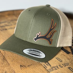 May include: Olive green and tan trucker hat with a deer antler patch. The hat features a curved brim, a mesh back, and a snapback closure. The patch is brown and black. The tag reads "The Authentic Snapback Yupoong Since 1974."