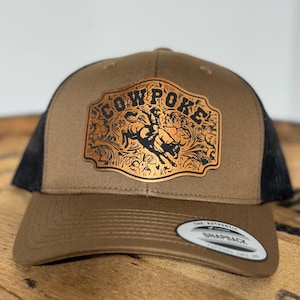 May include: A brown and black trucker hat with a leather patch that says "COWPOKE" in black letters with a gold background. The patch features a black silhouette of a cowboy riding a bucking bronco.
