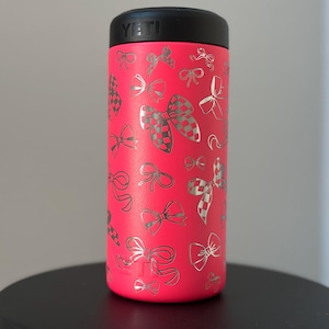 以下が含まれることがあります： 黒い蓋が付いたピンクのYETIランブラー。タンブラーは、銀色のリボンとチェック柄のリボンのパターンで覆われています。