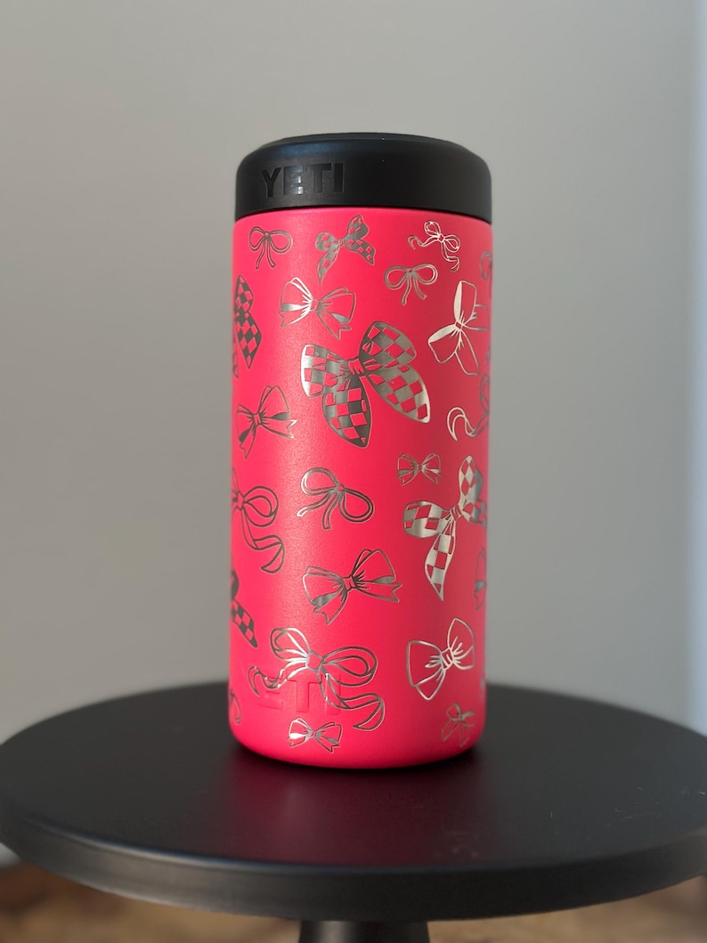 以下が含まれることがあります： 黒い蓋が付いたピンクのYETIランブラー。タンブラーは、銀色のリボンとチェック柄のリボンのパターンで覆われています。
