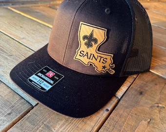 May include: A black trucker hat featuring a tan leather-like patch shaped like the state of Louisiana. The patch displays a black fleur-de-lis symbol above the text SAINTS and a small star. A sticker on the brim reads RICHARDSON 112 TRUCKER.