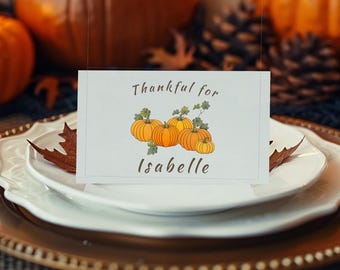 以下が含まれることがあります： 感謝祭のテーブルセッティング。キャンドル、カボチャ、そして「Thankful for Isabelle」と書かれた席札が置かれています。金色の縁取りのプレート、カトラリー、ワイングラスが並び、温かく居心地の良い雰囲気を作り出しています。
