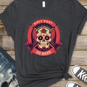 May include: Dark gray t-shirt featuring a colorful skull design with the text "Knit Fast Die Warm". The graphic includes a rose, wings, and knitting needles. The shirt is paired with blue jeans and sneakers.