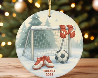 Op de afbeelding: Ronde kerstversiering met een voetbalthema. Het ontwerp toont een doel, bal, schoenen en rode handschoenen. De versiering heeft de naam "Isabella" en het jaar "2025" erop gedrukt. De achtergrond toont een besneeuwd landschap met dennenbomen.