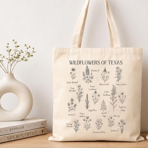 Könnte beinhalten: Eine cremefarbene Canvas-Tragetasche mit einem schwarzen botanischen Aufdruck mit dem Titel Wildflowers of Texas. Das Design zeigt verschiedene illustrierte Wildblumen, deren Namen in einer feinen Schreibschrift gedruckt sind, darunter Bluebonnet und Indian Paintbrush.