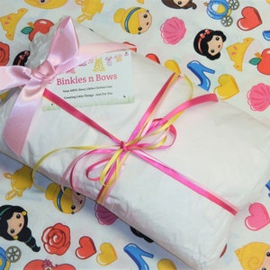 May include: A white gift wrapped in tissue paper with pink and yellow ribbons. The gift is sitting on a white background with a pattern of princess characters, hearts, apples, and crowns. The gift tag says "Binkies n Bows".