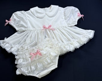 May include: A white dress with a ruffled hem and pink bows. The dress has short sleeves and a Peter Pan collar. It is paired with a white lace-trimmed bloomers with a pink bow.