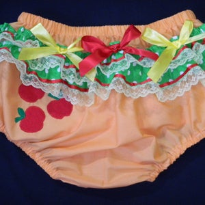 May include: Peach-coloured baby bloomers with an apple design. The bloomers have a ruffled trim with green polka dots, red ribbon, and white lace. Red and yellow bows adorn the front.