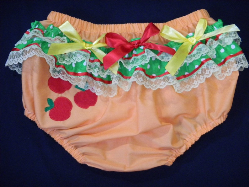 May include: Peach-coloured baby bloomers with an apple design. The bloomers have a ruffled trim with green polka dots, red ribbon, and white lace. Red and yellow bows adorn the front.