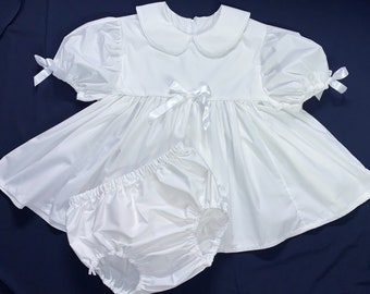 May include: A white dress with puffed sleeves and a Peter Pan collar, tied with a white satin bow at the waist. The dress is paired with a white cotton diaper cover.