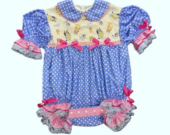 Può includere: Pagliaccetto per neonati a pois blu e bianchi, con colletto e maniche corte. Presenta un pannello color crema con personaggi dei cartoni animati, fiocchi rosa e bordi in pizzo. Il design è allegro e colorato.