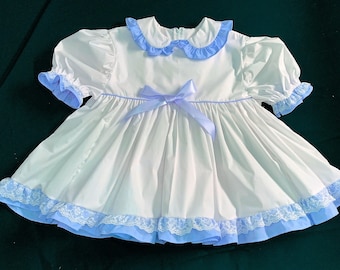 May include: White baby dress with light blue accents on the collar, sleeves, and hem. The dress has a light blue ribbon bow and lace trim. It features short, puffed sleeves and a ruffled collar. The dress is made of cotton.