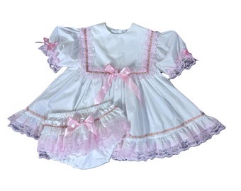 May include: A white dress and matching diaper cover set. The dress features a square neckline, short sleeves, and a full skirt, all trimmed with pink lace. Pink bows adorn the dress and diaper cover.