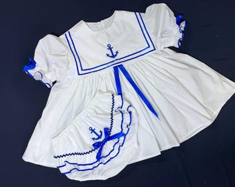 Può includere: Abito bianco da marinaio con bordi blu e un'ancora ricamata sul corpetto. L'abito ha un nastro blu legato in vita. L'abito è abbinato a pantaloncini bianchi con bordi blu e un'ancora ricamata sul davanti.