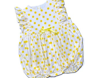 May include: White romper with yellow polka dots. The sleeveless garment has frilled sleeves and a yellow bow at the waist. The fabric is gathered at the bottom, creating a bubble shape. A zip runs down the back.