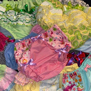 May include: A collection of colorful and patterned fabric baby bloomers with ruffles and bows. The bloomers are made of various fabrics, including gingham, floral, and polka dot.