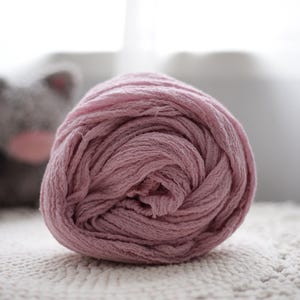 Puede incluir: Una pieza de tela de color rosa polvoriento enrollada firmemente que presenta una superficie texturizada y arrugada. El material está enrollado en forma de espiral circular, mostrando los pliegues superpuestos y el aspecto mate del tejido.
