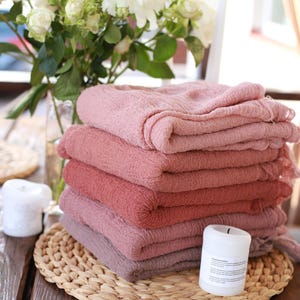 Puede incluir: Una pila de seis paños de tela doblados, en tonos rosados y malvas. Los paños están colocados sobre un salvamanteles tejido sobre una mesa de madera. También hay un jarrón con flores blancas y dos velas blancas.