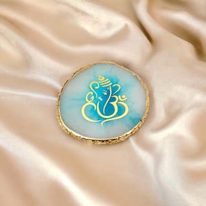 Op de afbeelding: Een ronde, lichtblauwe onderzetter met een goudkleurige rand en een goudkleurige illustratie van een godheid. De onderzetter is gemaakt van een doorschijnend materiaal en ligt op een satijnen oppervlak.