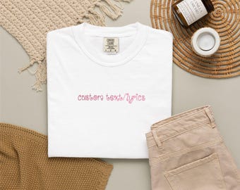 Puede incluir: Una camiseta blanca de manga corta con cuello redondo y una etiqueta de la marca Comfort Colors en el cuello interior. El centro del pecho presenta un texto bordado en rosa que dice custom text/lyrics en una fuente cursiva redondeada.
