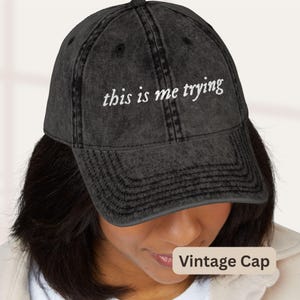 May include: A charcoal grey baseball cap with a distressed, washed-out finish. The front of the cap features the white embroidered text "this is me trying" in a cursive font. The cap has a structured crown, a curved brim, and stitched eyelets.